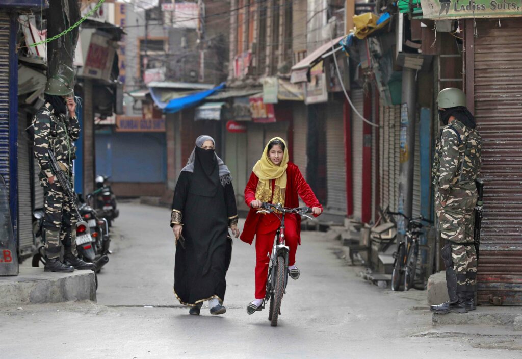 A Kashmir girl rides her bike past security personnel standing guard in front closed shops. Credit: Danish Ismail/Reuters